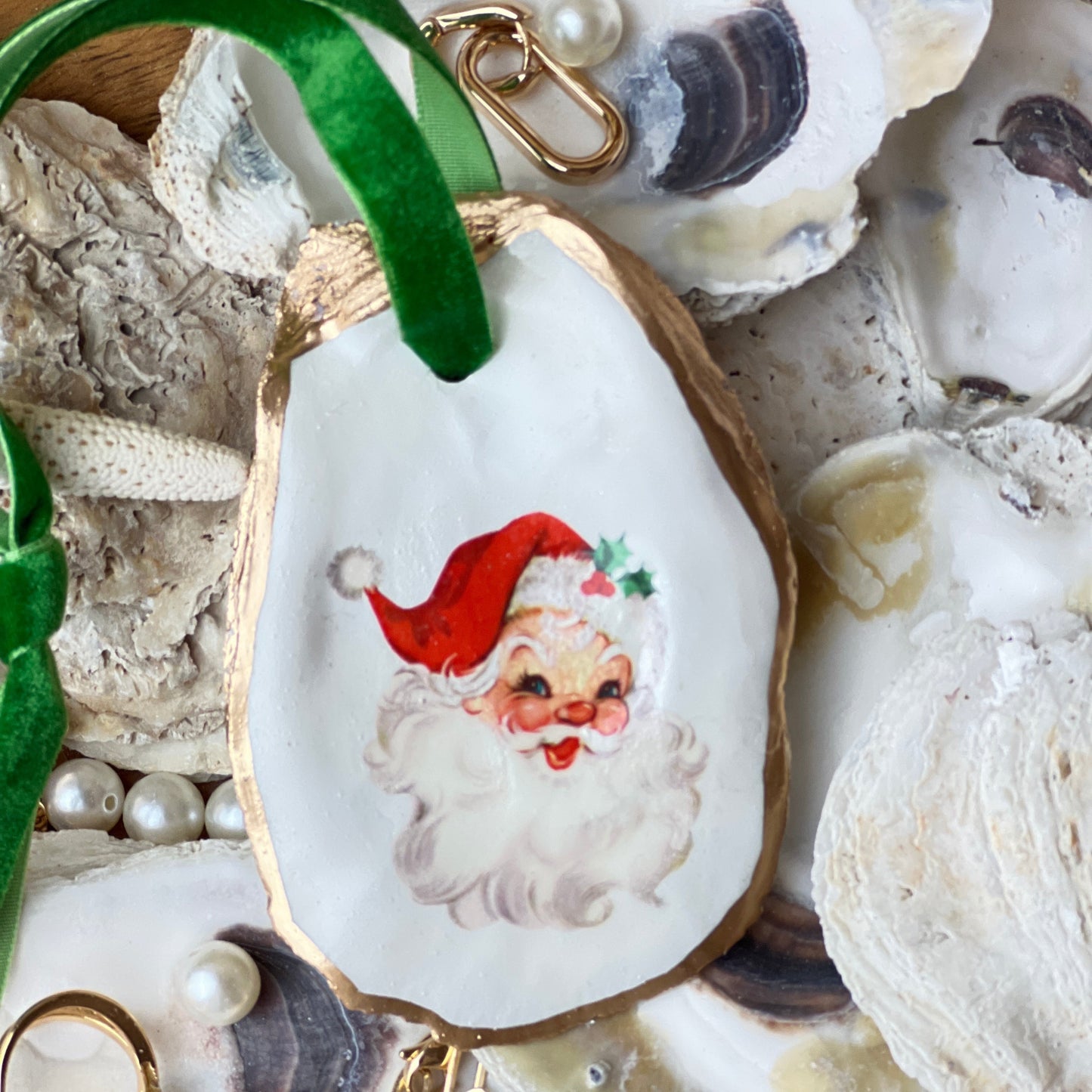 A hand-decorated retro Santa ornament on a natural oyster shell with a red Santa Claus design, accented with liquid gold leaf and a green velvet ribbon.