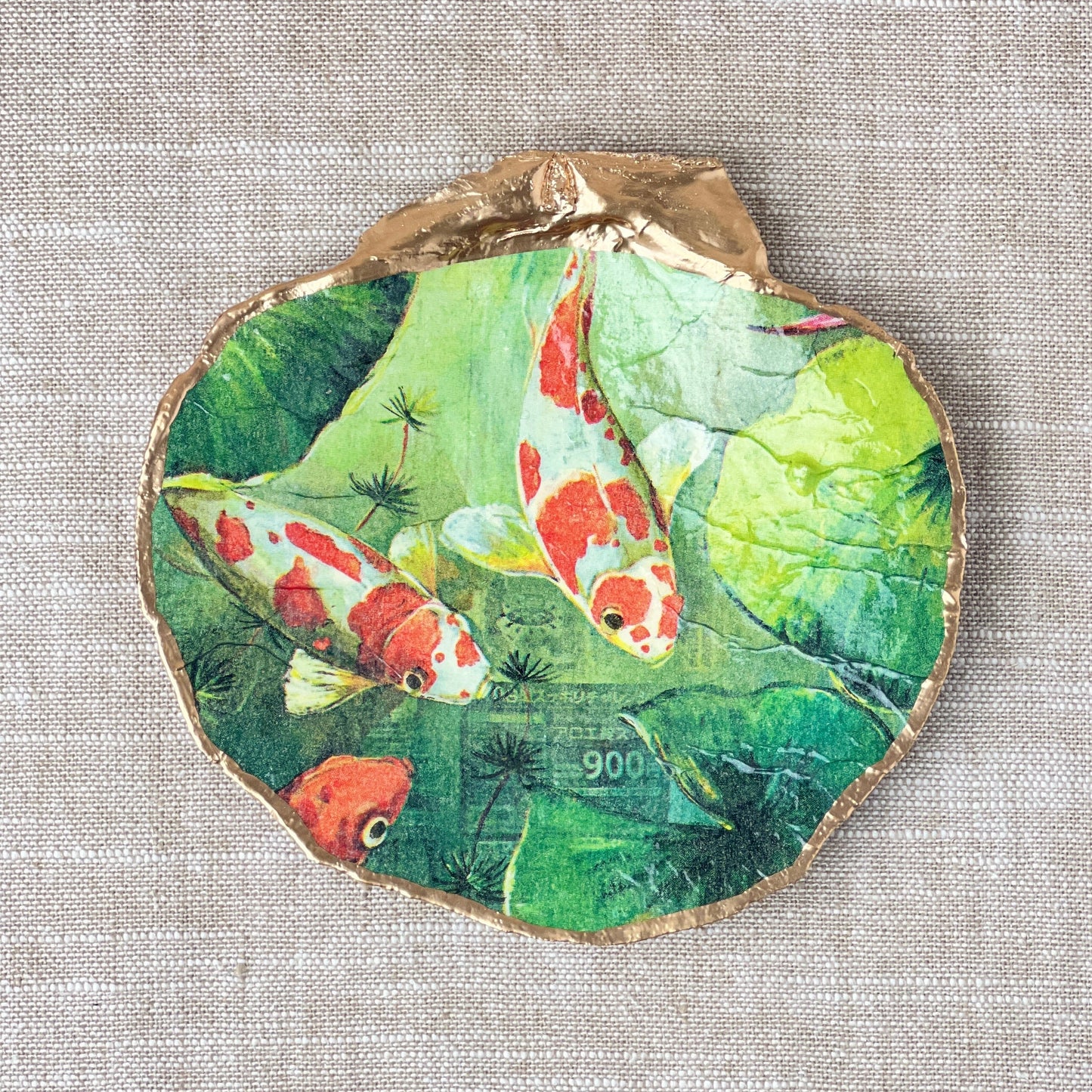 A scallop shell ring dish with a green and orange koi pond design, displayed flat on a neutral linen cloth.