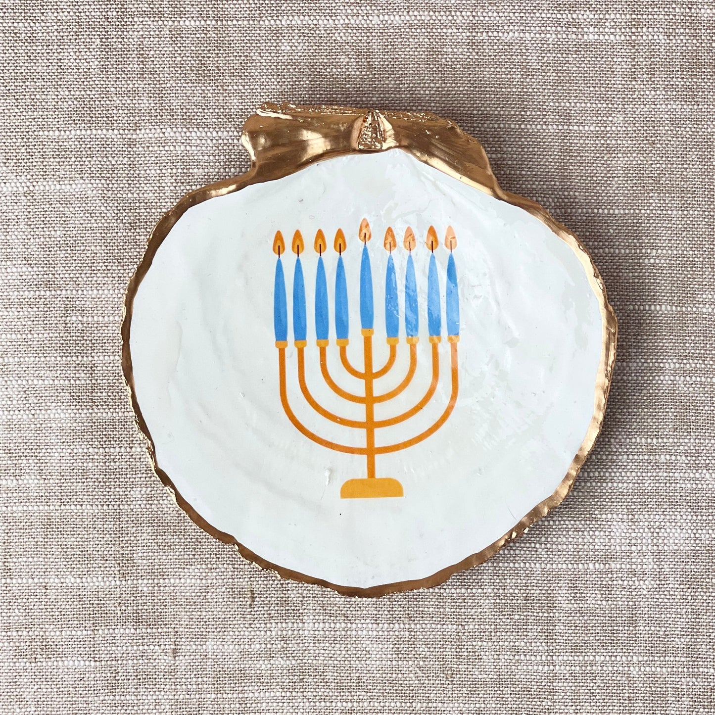A scallop shell with a hand-decorated Menorah design in blue and orange colors, displayed flat on a neutral background.