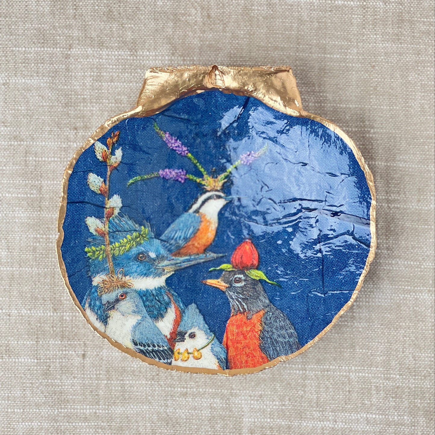 A scallop shell ring dish with a navy background featuring colorful bird designs, displayed flat n a beige surface.