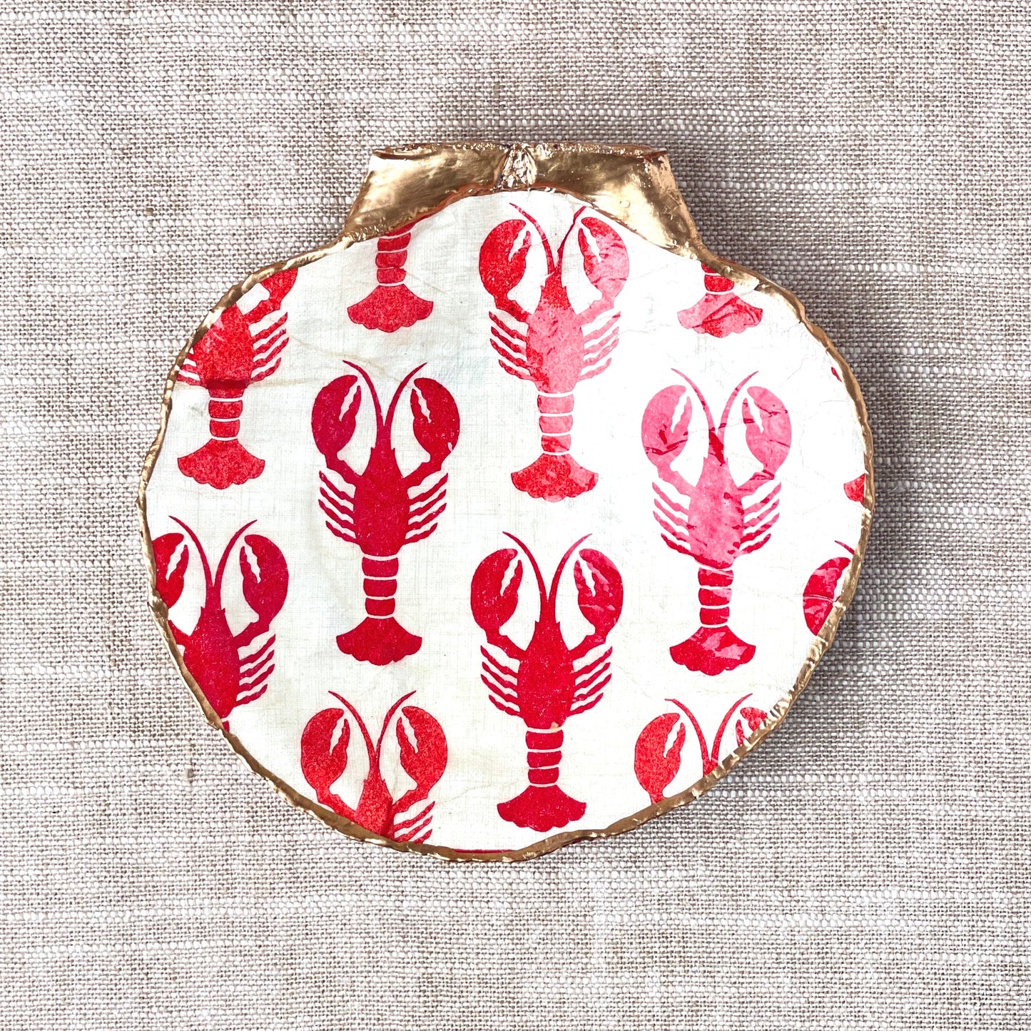 A scallop shell ring dish with a hand-decorated lobster design in red on a white background placed on a tan linen cloth background.