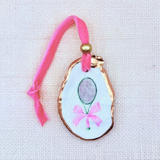 Decorative Christmas oyster ornament with pink tennis racket and bow design on a light background.