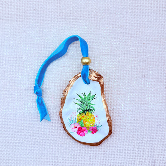 Decorative oyster shell with a pineapple and floral design, tied with a blue ribbon on a neutral background.