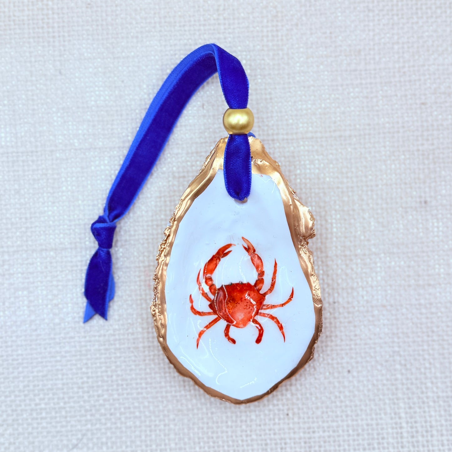 Decorative oyster ornament with a red crab design on a white background, attached to a blue ribbon.