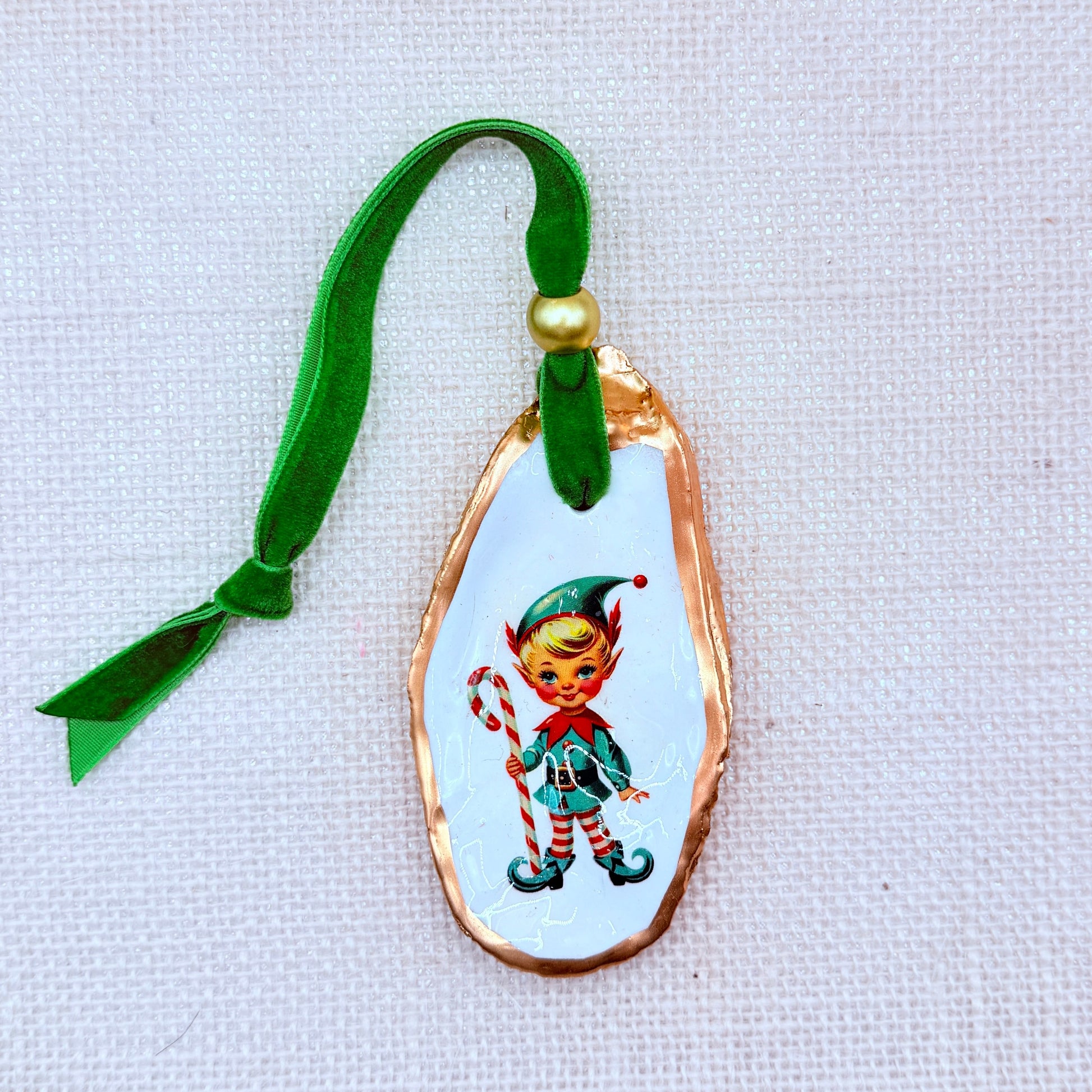Decorative oyster ornament of an elf design and green ribbon on a light fabric background.