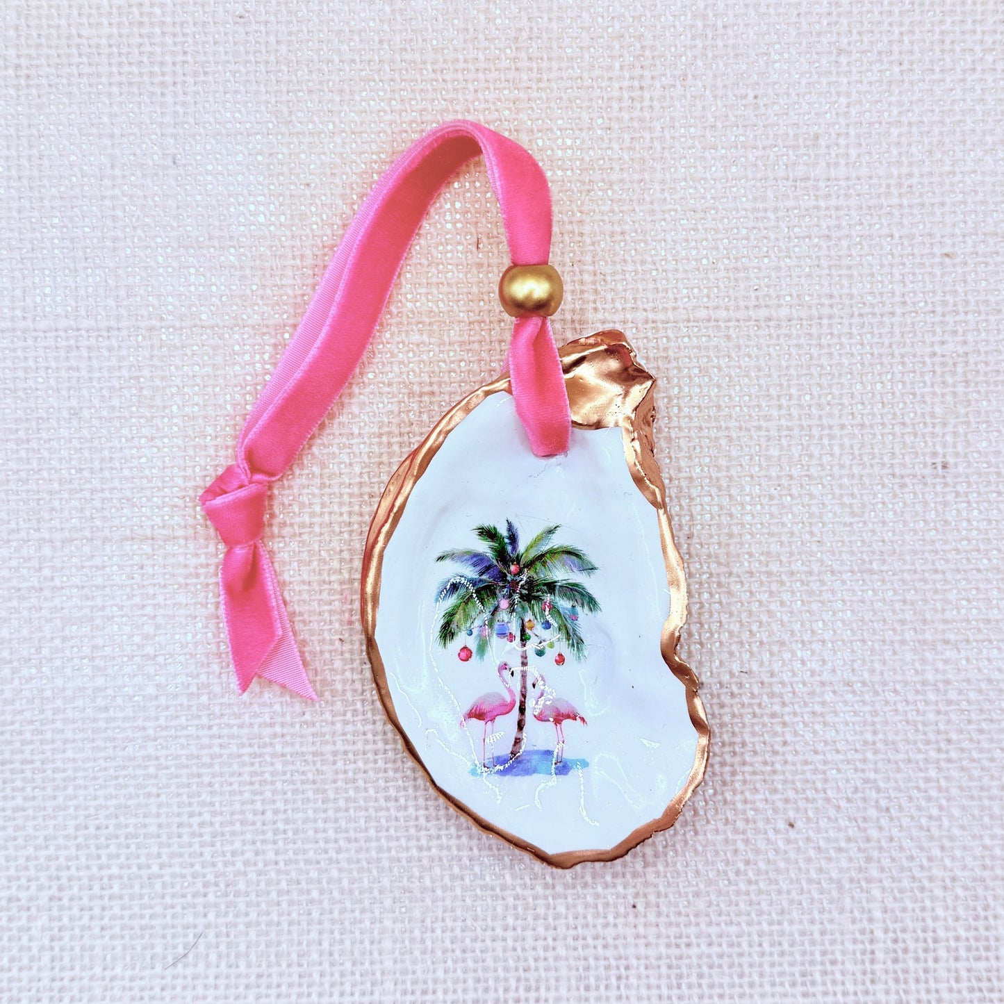 Decorative oyster ornament with palm tree design, flamingos, and pink ribbon on a light background.