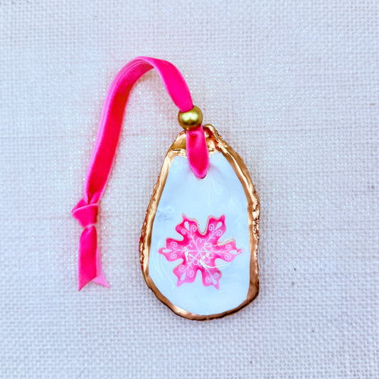 oval-shaped ornament with pink snowflake design  ON AN OYSTER SHELL WITH gold trim on a pink ribbon against a neutra  background.