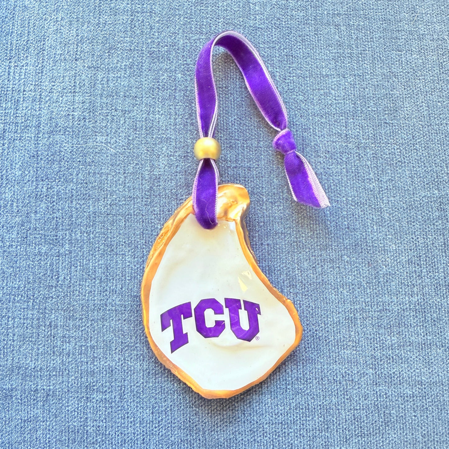 Decorative oyster Christmas ornament with 'TCU' on a purple ribbon against a blue textured background.
