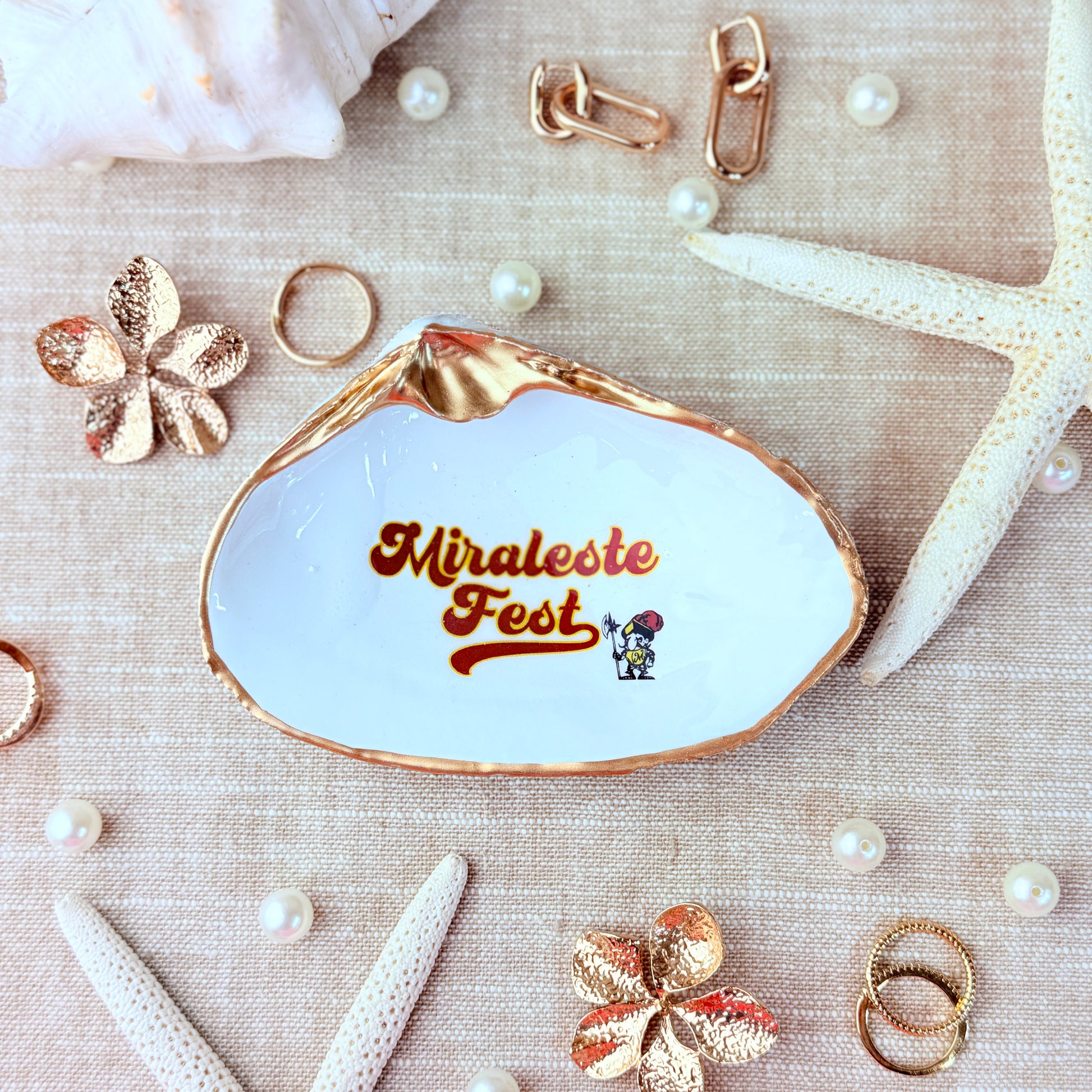 Clam shell-shaped jewelry dish with 'Miraleste Fest' text surrounded by decorative items on a textured beige surface.