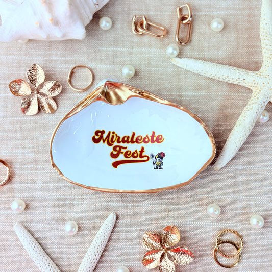 Clam shell-shaped jewelry dish with 'Miraleste Fest' text surrounded by decorative items on a textured beige surface.