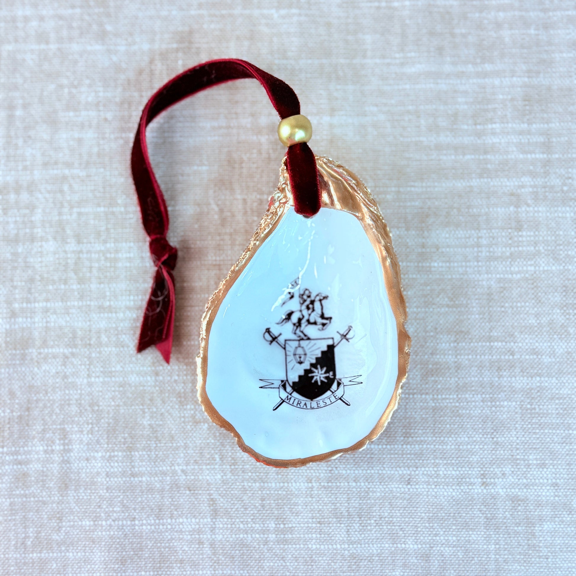 Decorative ornament with the Miraleste High School Crest design on a light white background, hanging from a burgundy ribbon.