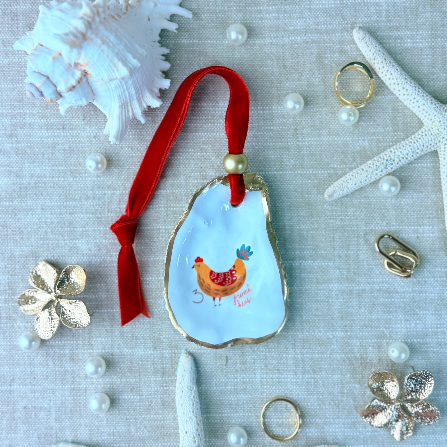 Decorative oyster shell with a red ribbon and chicken illustration on a textured surface with shells and pearls.